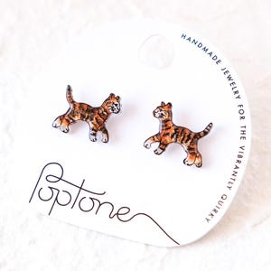 May include: A pair of cat-shaped stud earrings. The earrings feature a brown and orange tabby cat design with black outlines. The earrings are on a white card with the word "Poptone" and the text "Handmade Jewelry for the Vibrantly Quirky".