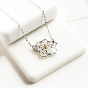 May include: A silver chain necklace with a delicate white flower pendant. The flower has a yellow center and is outlined in black.