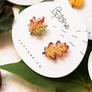 Puede incluir: Un par de pendientes de hojas de oto&ntilde;o, uno es una hoja de arce y el otro es una hoja de roble. Las hojas est&aacute;n pintadas en tonos de naranja, amarillo y verde. Los pendientes est&aacute;n en una tarjeta blanca con el texto "Poptone.co" impreso en ella.