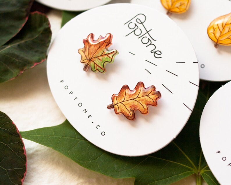 Puede incluir: Un par de pendientes de hojas de oto&ntilde;o, uno es una hoja de arce y el otro es una hoja de roble. Las hojas est&aacute;n pintadas en tonos de naranja, amarillo y verde. Los pendientes est&aacute;n en una tarjeta blanca con el texto "Poptone.co" impreso en ella.
