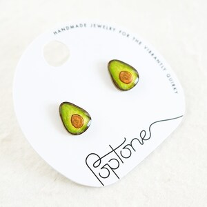 Puede incluir: Un par de pendientes con forma de aguacate con un dise&ntilde;o verde y marr&oacute;n. Los pendientes est&aacute;n en una tarjeta blanca con el texto "Handmade Jewelry for the Vibrantly Quirky" y "Poptones".