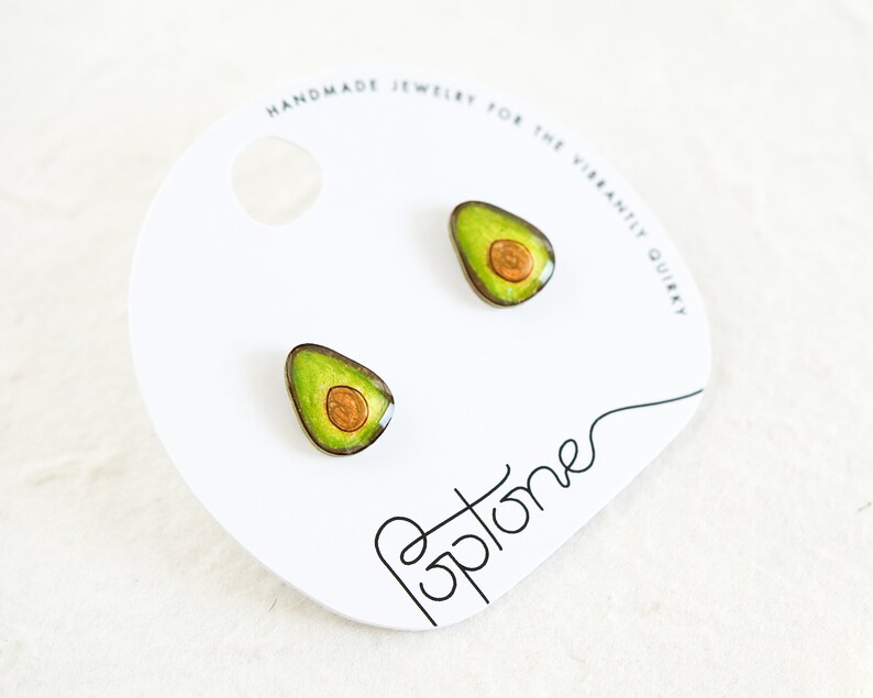 Puede incluir: Un par de pendientes con forma de aguacate con un dise&ntilde;o verde y marr&oacute;n. Los pendientes est&aacute;n en una tarjeta blanca con el texto "Handmade Jewelry for the Vibrantly Quirky" y "Poptones".