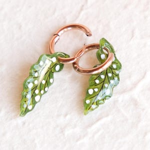 May include: A pair of rose gold hoop earrings with green leaf charms. The charms have a white polka dot pattern.
