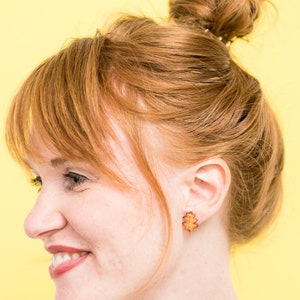 Puede incluir: Una mujer con el pelo rojo en un mo&ntilde;o desordenado lleva un par de peque&ntilde;os pendientes de hoja de oro. Los pendientes tienen forma de hojas de roble y son de color marr&oacute;n rojizo.