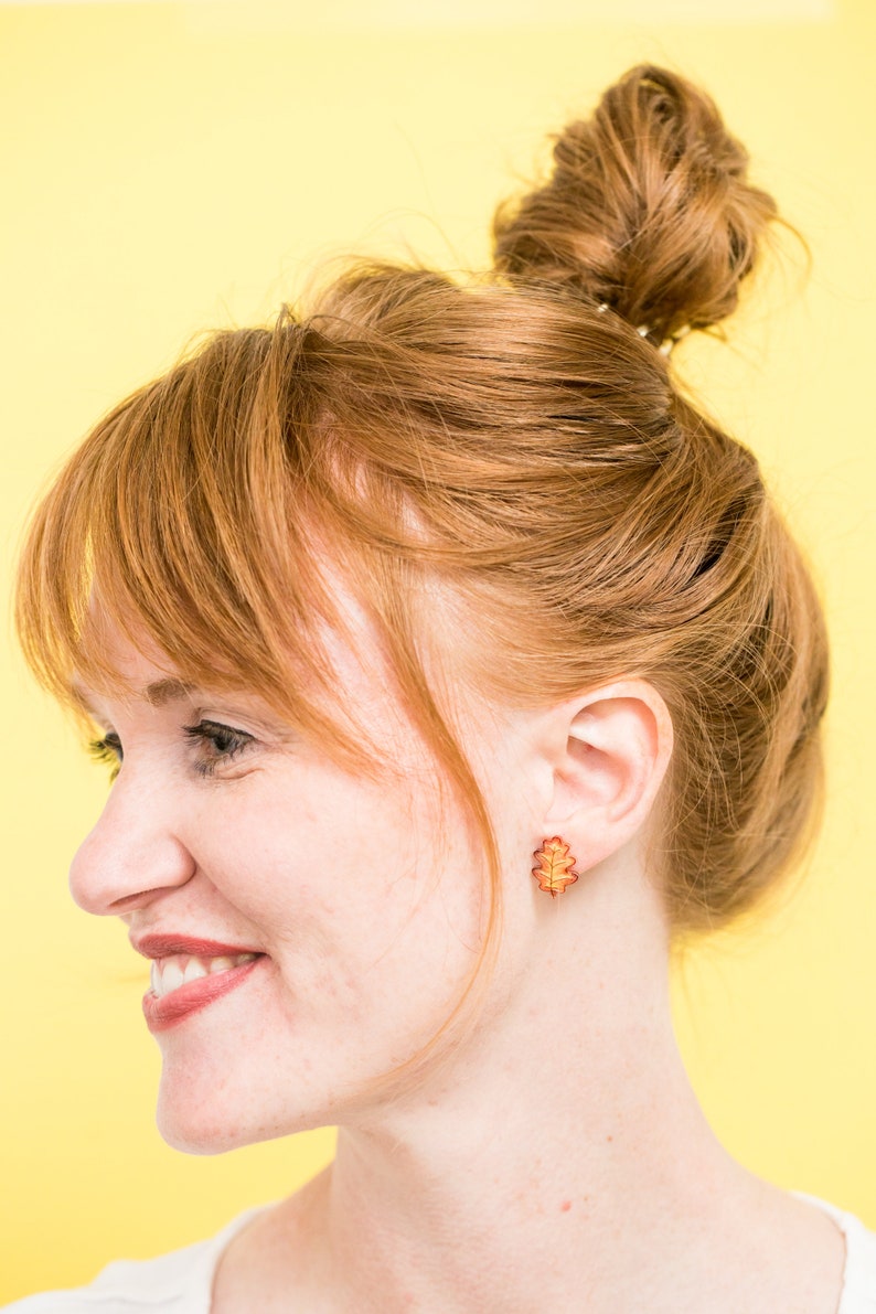 Puede incluir: Una mujer con el pelo rojo en un mo&ntilde;o desordenado lleva un par de peque&ntilde;os pendientes de hoja de oro. Los pendientes tienen forma de hojas de roble y son de color marr&oacute;n rojizo.