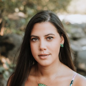 May include: A woman with long dark hair wearing a pink and green floral print top with a green leaf necklace and matching earrings.