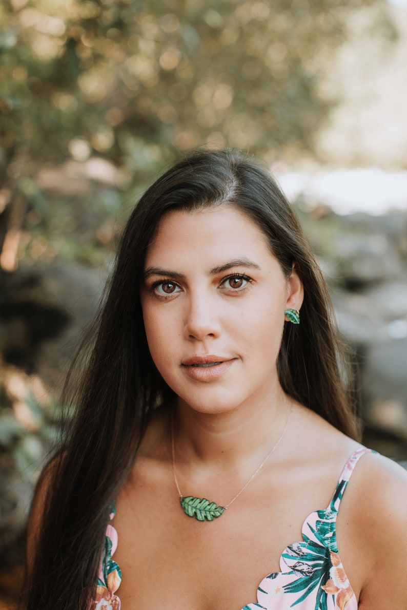 May include: A woman with long dark hair wearing a pink and green floral print top with a green leaf necklace and matching earrings.