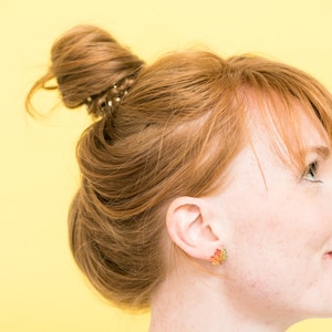 Puede incluir: Una mujer con el pelo rojo recogido en un mo&ntilde;o desordenado con una goma de pelo marr&oacute;n. Lleva unos peque&ntilde;os pendientes de oro en forma de hojas de arce con detalles en rojo, amarillo y naranja.