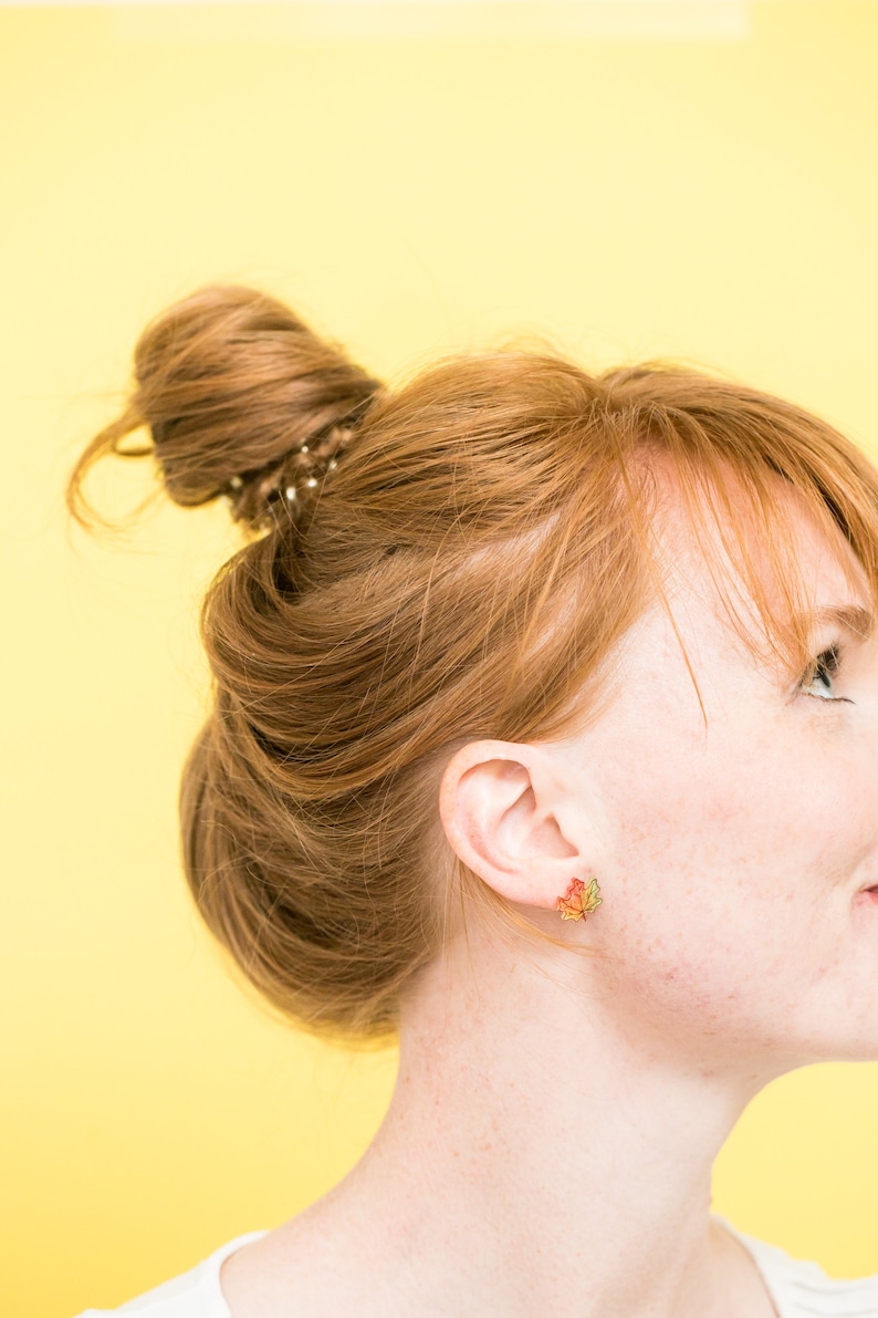 Puede incluir: Una mujer con el pelo rojo recogido en un mo&ntilde;o desordenado con una goma de pelo marr&oacute;n. Lleva unos peque&ntilde;os pendientes de oro en forma de hojas de arce con detalles en rojo, amarillo y naranja.