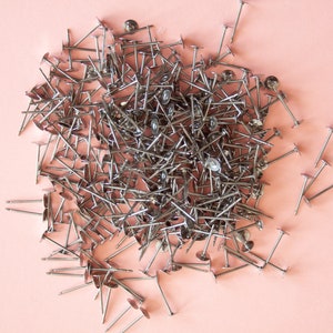 May include: A large pile of silver-toned metal tacks with flat heads and pointed ends, scattered on a light pink surface.