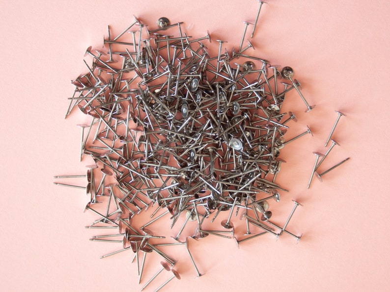 May include: A large pile of silver-toned metal tacks with flat heads and pointed ends, scattered on a light pink surface.