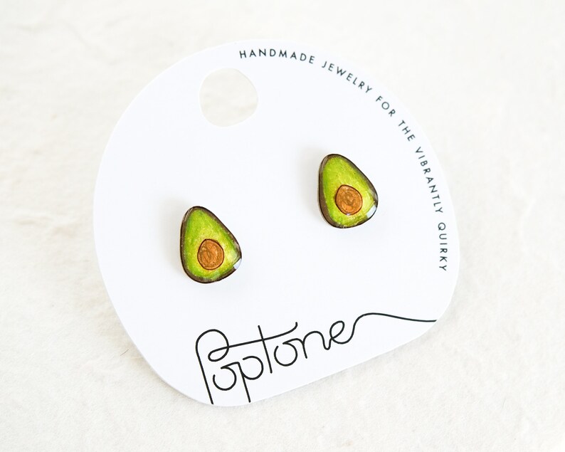 Puede incluir: Un par de pendientes con forma de aguacate con un dise&ntilde;o verde y marr&oacute;n. Los pendientes est&aacute;n en una tarjeta blanca con el texto "HANDMADE JEWELRY FOR THE VIBRANTLY QUIRKY" y la palabra "optone" escrita en cursiva.