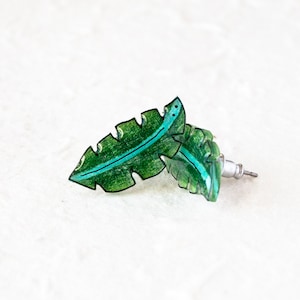 May include: A pair of green leaf-shaped earrings with a black outline and a turquoise line down the center. The earrings are made of resin and have a silver post.
