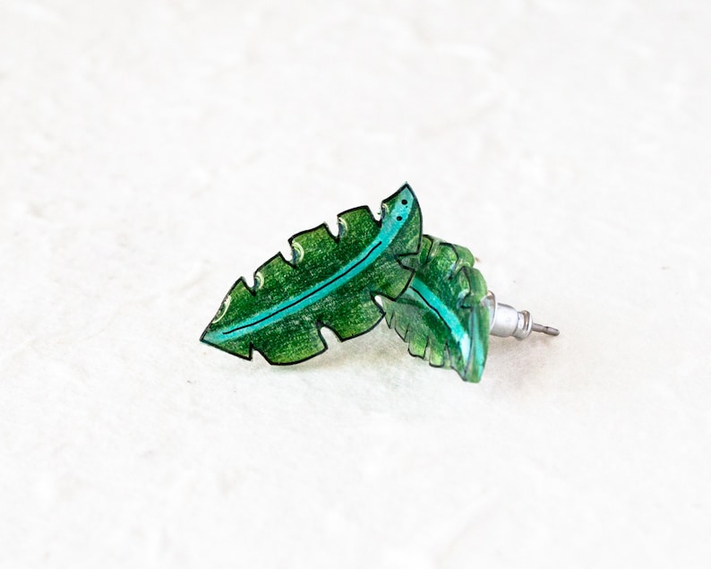 May include: A pair of green leaf-shaped earrings with a black outline and a turquoise line down the center. The earrings are made of resin and have a silver post.