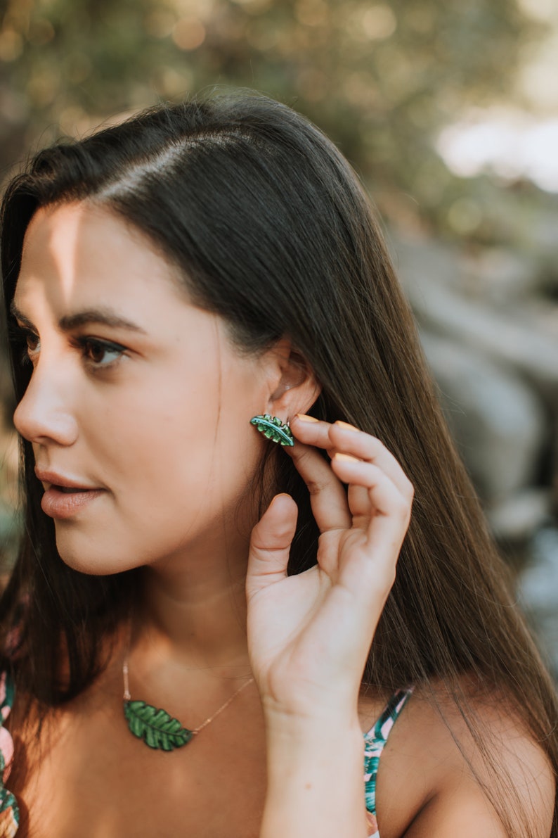 May include: A woman wearing a green leaf necklace and earrings. The earrings are small and shaped like leaves. The necklace is a delicate chain with a larger leaf pendant.