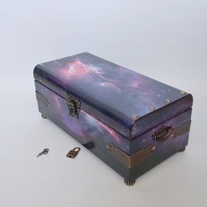 Può includere: Una scatola di legno con un coperchio stampato con una galassia, dotata di serratura e chiave. La scatola ha dettagli in ottone e poggia su quattro piccoli piedini.