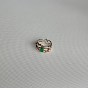 May include: A silver and rose gold ring with a green gemstone set in the center. The ring has a unique, organic design.