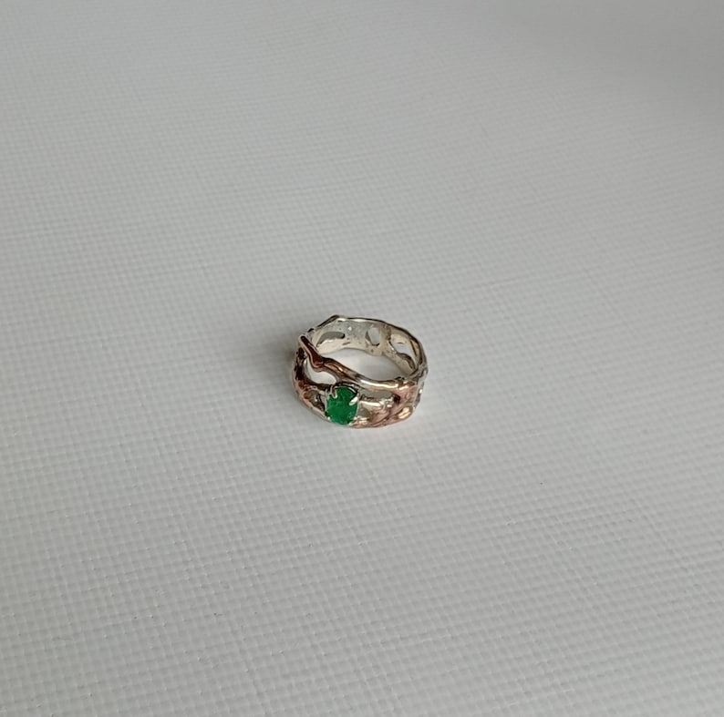 May include: A silver and rose gold ring with a green gemstone set in the center. The ring has a unique, organic design.