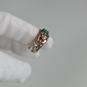 May include: A silver ring with a green gemstone set in a delicate, intricate design. The ring is made of two different metals, with a silver band and a gold accent.