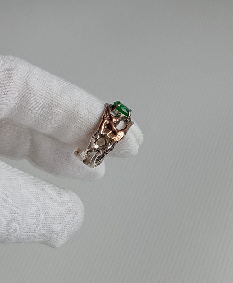 May include: A silver ring with a green gemstone set in a delicate, intricate design. The ring is made of two different metals, with a silver band and a gold accent.