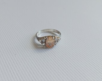 May include: A silver ring with a large oval-shaped peach-colored gemstone surrounded by smaller clear gemstones.