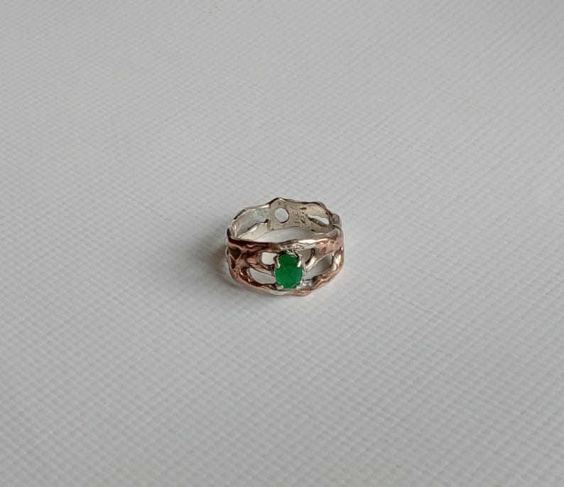 May include: A silver and copper ring with a green gemstone set in the center. The ring has a unique, organic design.