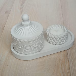 Puede incluir: Bandeja de cerámica blanca con un recipiente decorativo con tapa y un pequeño portavelas. El recipiente tiene un diseño en relieve y el portavelas tiene forma de cuerda.