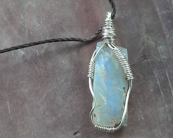 May include: A silver wire wrapped pendant featuring a light blue, opaque, irregular shaped stone. The pendant is hanging from a black cord.