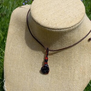 May include: A black gemstone pendant with red wire wrapping hangs from a brown leather cord necklace.