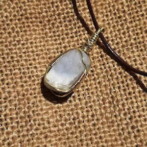 Puede incluir: Un delicado alambre de oro envuelto alrededor de un colgante de piedra preciosa transparente e iridiscente. El colgante cuelga de un cordón de cuero marrón.