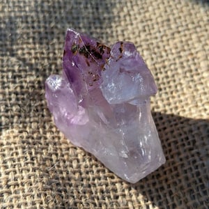 Puede incluir: Un grupo de cristales de amatista, que muestra un degradado de colores desde el púrpura intenso hasta el blanco translúcido. La formación natural tiene una superficie rugosa y texturizada con pequeñas inclusiones oscuras. Los cristales se exhiben sobre una superficie texturizada de tono neutro.