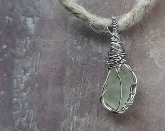 May include: A handmade pendant featuring a pale green stone wrapped in silver-colored wire. The pendant hangs from a natural-colored, woven cord. The background is a textured, muted brown surface.
