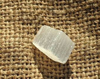 Può includere: Un cristallo di selenite bianco trasparente con forma rettangolare. Il cristallo è su una superficie di juta marrone.