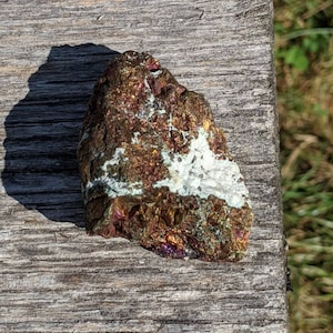 May include: A close-up of a rough, iridescent, metallic rock with a white vein running through it. The rock is sitting on a weathered wooden surface.