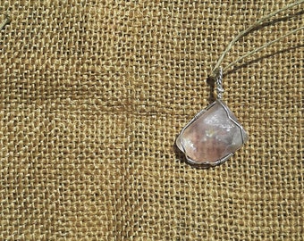 May include: A pink and white crystal pendant wrapped in silver wire, hanging from a brown string on a hessian background.