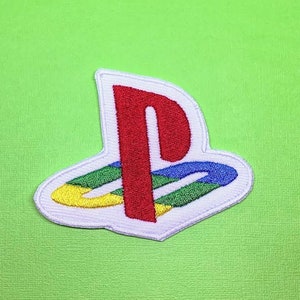 Può includere: Toppa ricamata con il logo PlayStation. La toppa ha un bordo bianco, una forma a "P" rossa e un design multicolore verde, blu e giallo. Lo sfondo è verde lime.