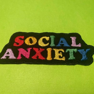 May include: A black embroidered patch with the words "SOCIAL ANXIETY" in colorful, stitched letters. The text is in a rainbow of colors, including red, yellow, pink, purple, green, and blue. The patch is on a bright green background.