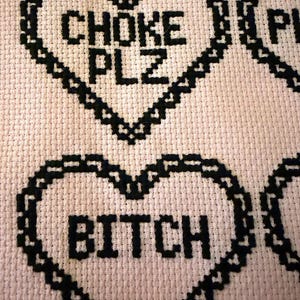 Puede incluir: Primer plano de un dise&ntilde;o de punto de cruz sobre una tela rosa claro. El dise&ntilde;o presenta corazones cosidos en negro con las palabras "CHOKE PLZ" y "BITCH" en el centro de los corazones. La costura es detallada y la tela tiene un aspecto texturizado.