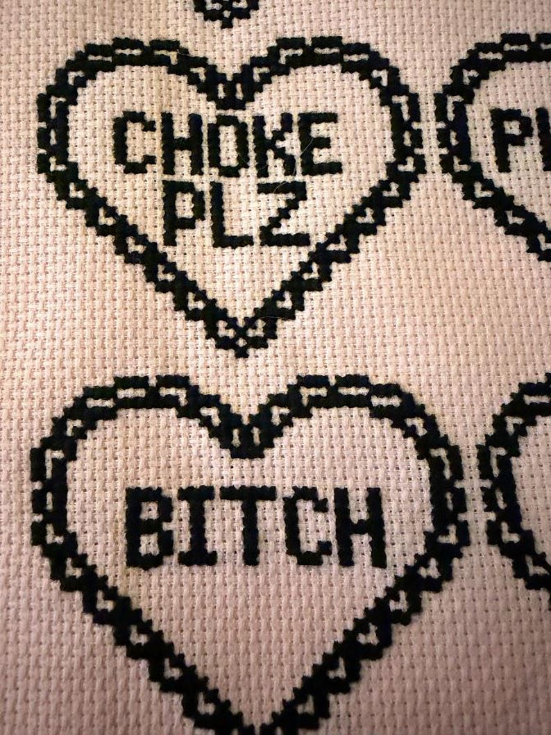 Puede incluir: Primer plano de un dise&ntilde;o de punto de cruz sobre una tela rosa claro. El dise&ntilde;o presenta corazones cosidos en negro con las palabras "CHOKE PLZ" y "BITCH" en el centro de los corazones. La costura es detallada y la tela tiene un aspecto texturizado.