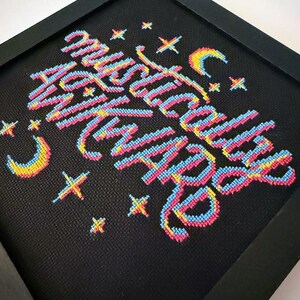 May include: Framed cross-stitch art with the words "Mystically With You" in colorful thread. The text is in a cursive style with pink, blue, and yellow hues. The black frame surrounds the artwork, which also features stars and crescent moons.