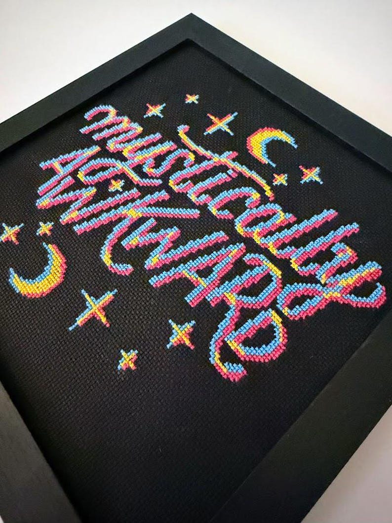 May include: Framed cross-stitch art with the words "Mystically With You" in colorful thread. The text is in a cursive style with pink, blue, and yellow hues. The black frame surrounds the artwork, which also features stars and crescent moons.