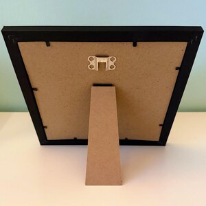 May include: A black picture frame with a cardboard backing and a brown easel stand. The frame is rectangular and has a simple, modern design. The easel stand is attached to the back of the frame.