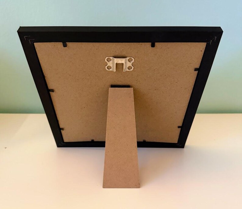 May include: A black picture frame with a cardboard backing and a brown easel stand. The frame is rectangular and has a simple, modern design. The easel stand is attached to the back of the frame.