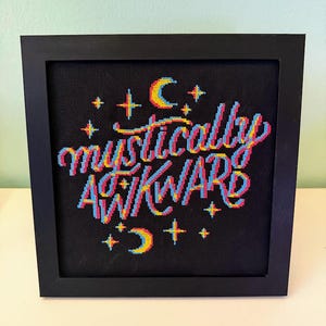 May include: Cross-stitch art in a black frame. The artwork features the words "mystically awkward" in colorful thread, with a 3D effect. Stars and crescent moons surround the text on a black background. The frame is square and made of black wood.