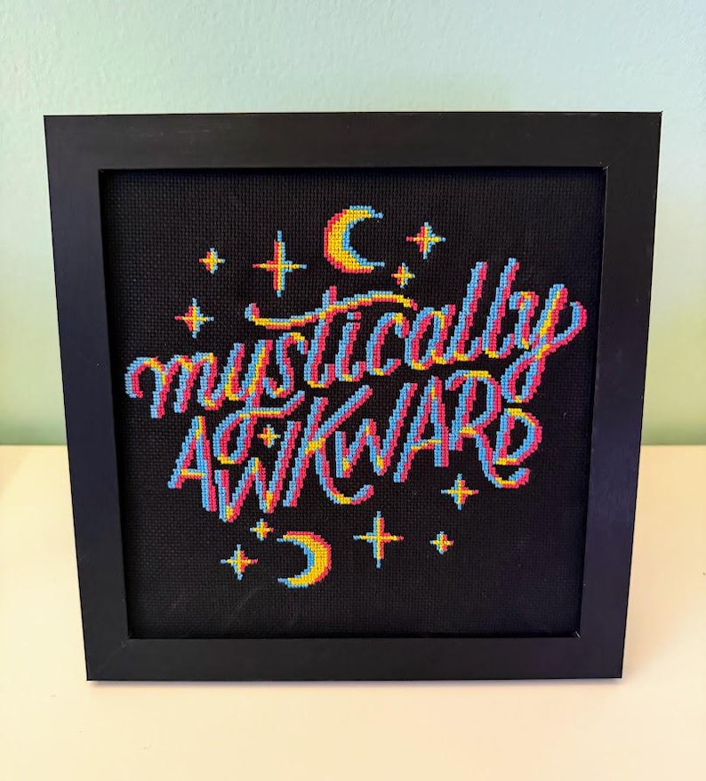 May include: Cross-stitch art in a black frame. The artwork features the words "mystically awkward" in colorful thread, with a 3D effect. Stars and crescent moons surround the text on a black background. The frame is square and made of black wood.