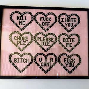 Puede incluir: Arte de punto de cruz enmarcado con un fondo rosa y dise&ntilde;os en forma de coraz&oacute;n negros. Cada coraz&oacute;n contiene un mensaje de texto en negrita, en negro, incluyendo frases como "KILL ME", "FUCK OFF" e "I HATE YOU". La obra de arte est&aacute; encerrada en un marco negro.