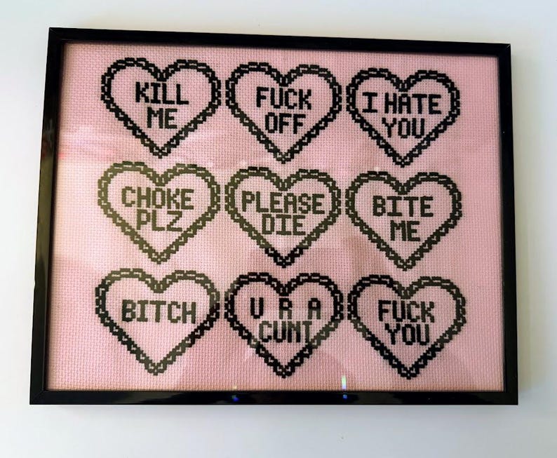 Puede incluir: Arte de punto de cruz enmarcado con un fondo rosa y dise&ntilde;os en forma de coraz&oacute;n negros. Cada coraz&oacute;n contiene un mensaje de texto en negrita, en negro, incluyendo frases como "KILL ME", "FUCK OFF" e "I HATE YOU". La obra de arte est&aacute; encerrada en un marco negro.