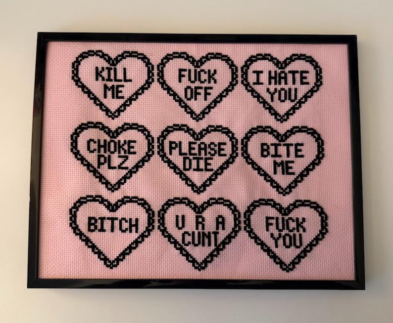Puede incluir: Una obra de arte de punto de cruz enmarcada con un fondo rosa. Nueve dise&ntilde;os negros en forma de coraz&oacute;n presentan varias frases, incluyendo "KILL ME", "FUCK OFF" y "I HATE YOU". La obra est&aacute; enmarcada en un marco negro.