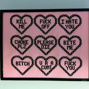 Puede incluir: Una obra de arte en punto de cruz enmarcada con un fondo rosa. Nueve formas de coraz&oacute;n negras est&aacute;n dispuestas en una cuadr&iacute;cula, cada una con una frase corta y provocativa en may&uacute;sculas, como "KILL ME" y "FUCK YOU". El marco es negro.