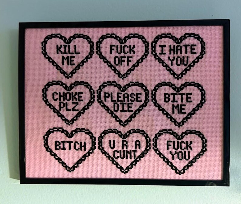 Puede incluir: Una obra de arte en punto de cruz enmarcada con un fondo rosa. Nueve formas de coraz&oacute;n negras est&aacute;n dispuestas en una cuadr&iacute;cula, cada una con una frase corta y provocativa en may&uacute;sculas, como "KILL ME" y "FUCK YOU". El marco es negro.